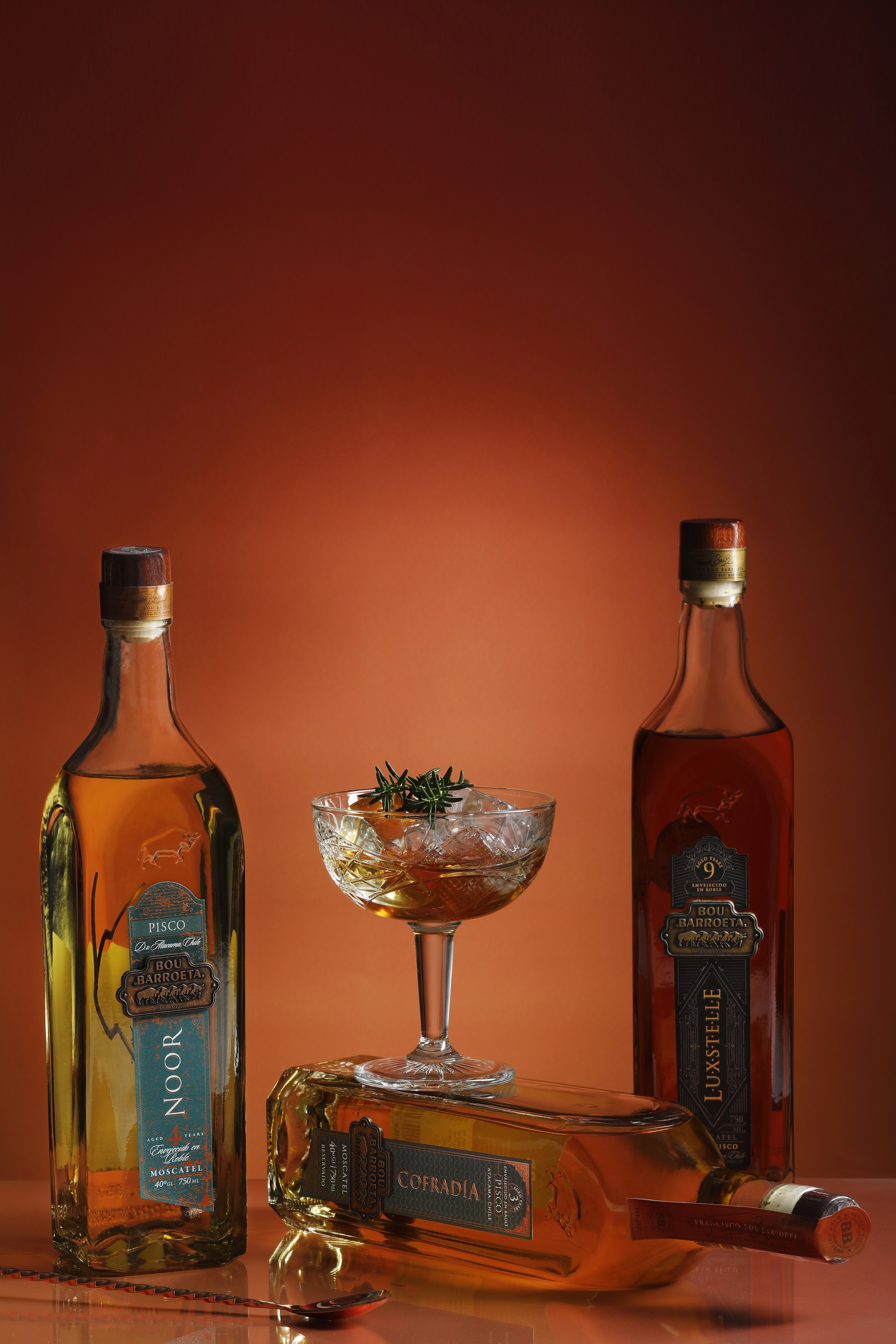 Dos botellas añejadas Bou Barroeta junto a una copa y una botella horizontal, sobre un fondo cobrizo.