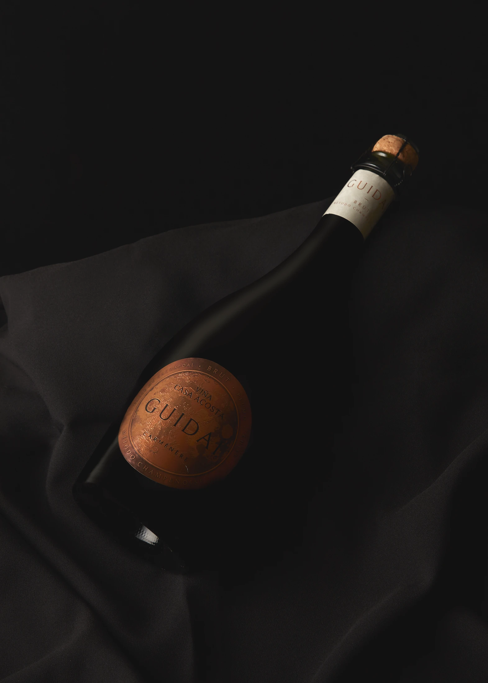 Botella Guidaí sobre seda negra, con el medallón de cobre en relieve como único punto de luz.