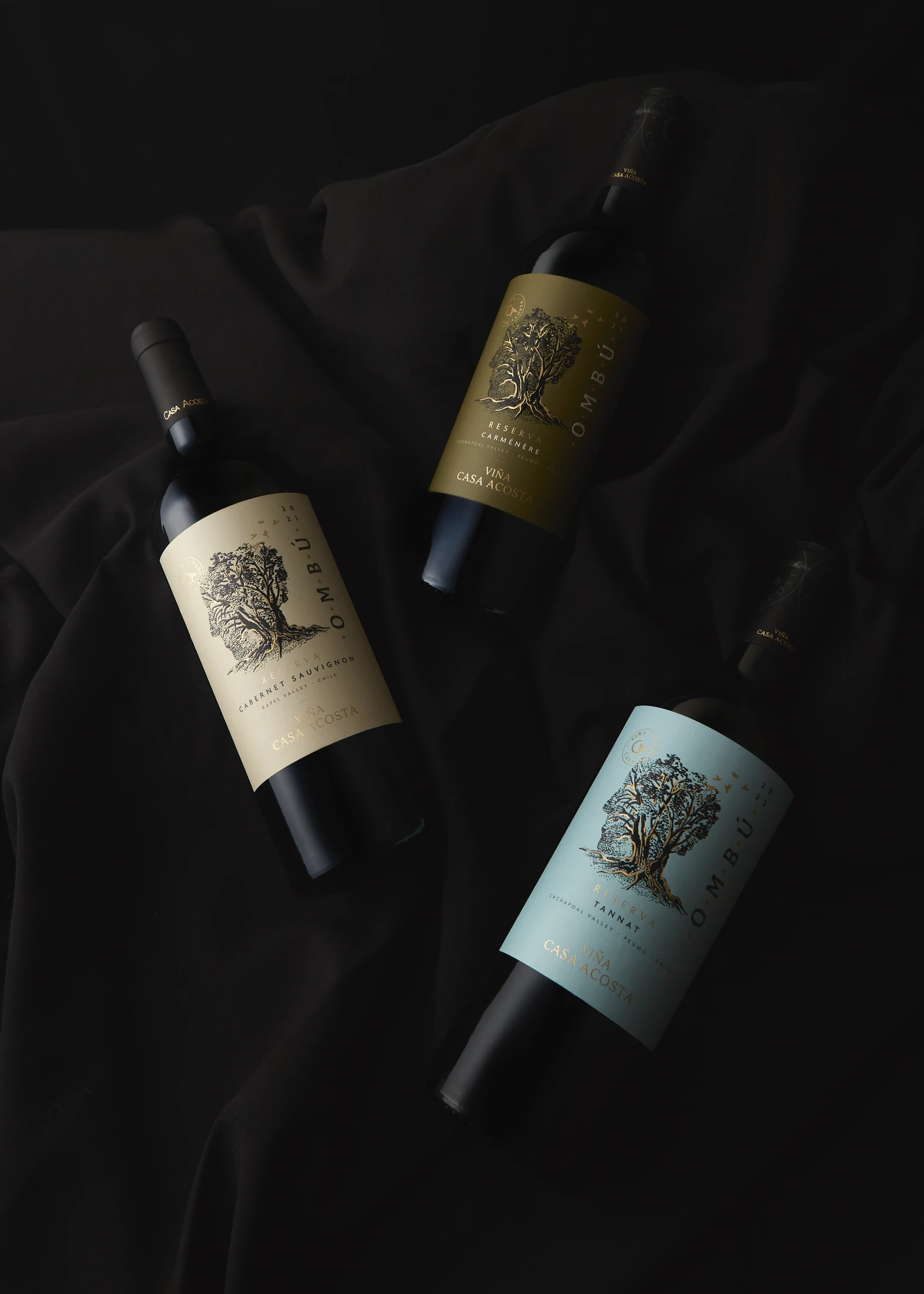 Tres botellas Ombú —Cabernet Sauvignon, Carménère y Tannat— sobre seda negra, con el mismo grabado del árbol en tres papeles distintos.