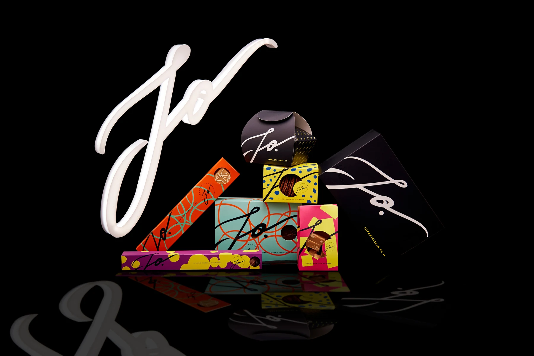 Sistema de packaging JO sobre fondo negro: cajas y estuches en verde agua, naranja, fucsia, amarillo y negro junto al logotipo caligráfico en volumen.