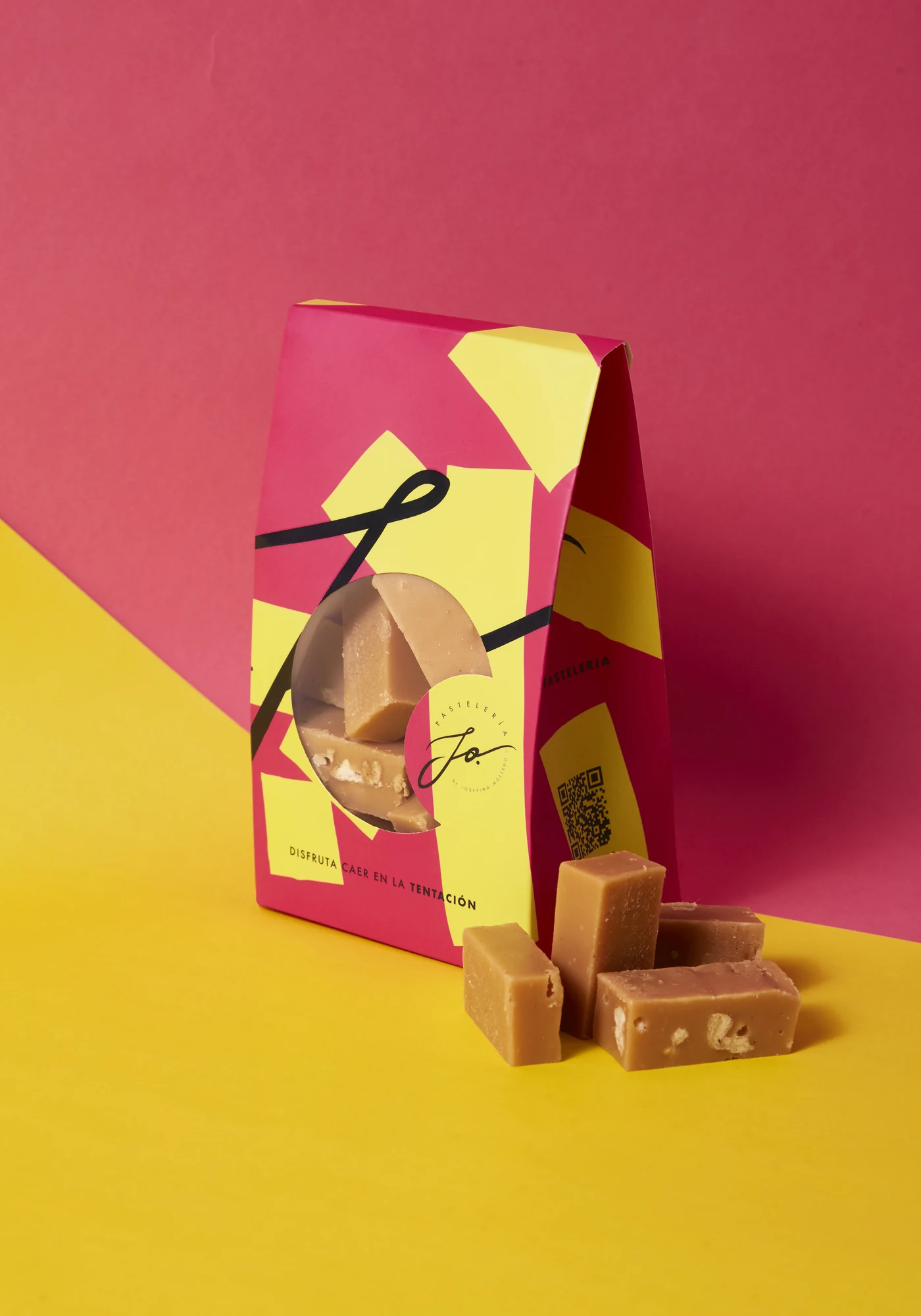 Estuche vertical JO en fucsia y amarillo con ventana troquelada y lazo negro, junto a calugas de manjar sobre un fondo bicolor.