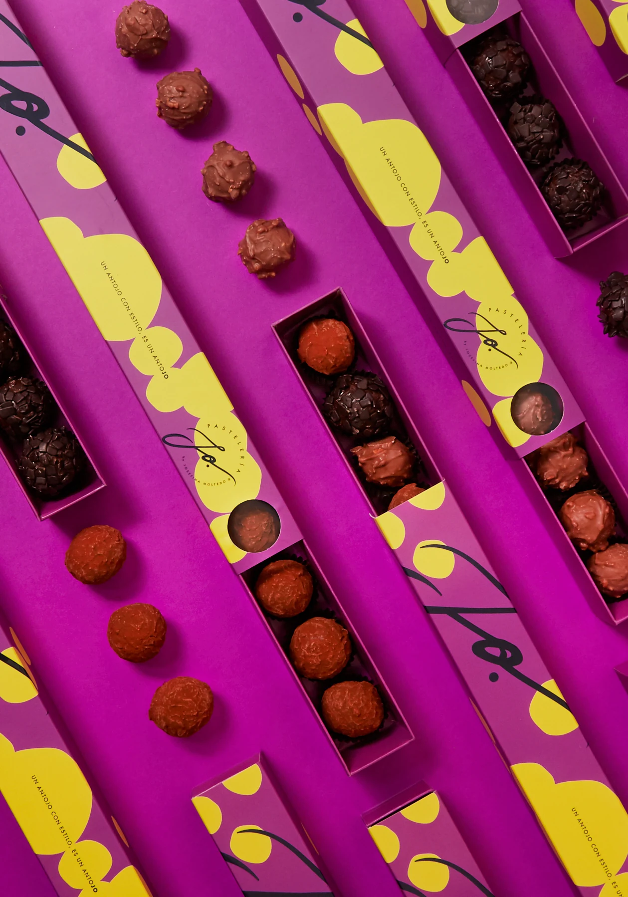 Estuches alargados JO en magenta con lunares amarillos, abiertos para mostrar trufas de chocolate de distintos sabores.