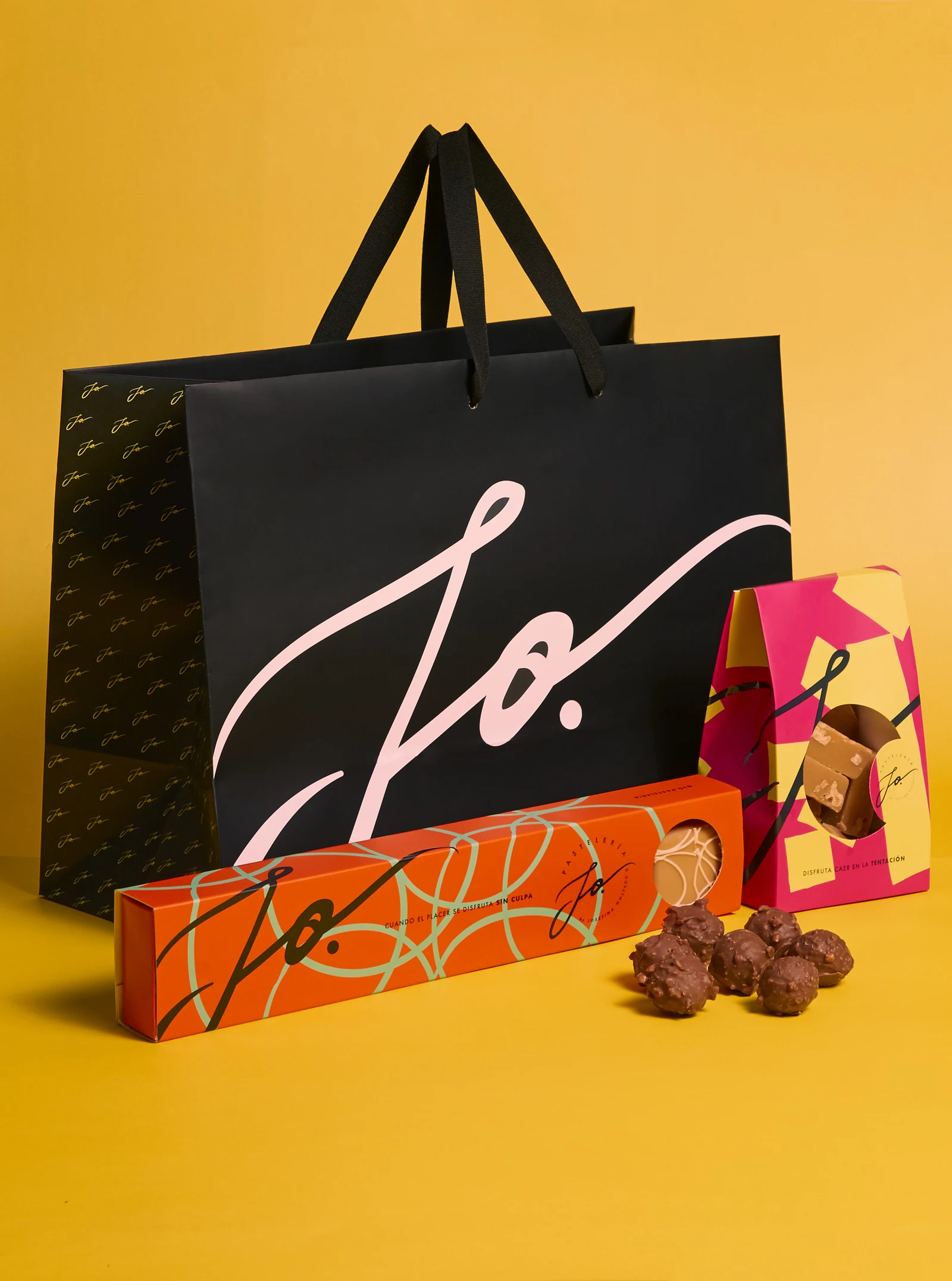 Sistema de marca JO sobre fondo amarillo: bolsa de regalo negra con la caligrafía en rosa, estuche alargado naranja y estuche fucsia junto a trufas de chocolate.