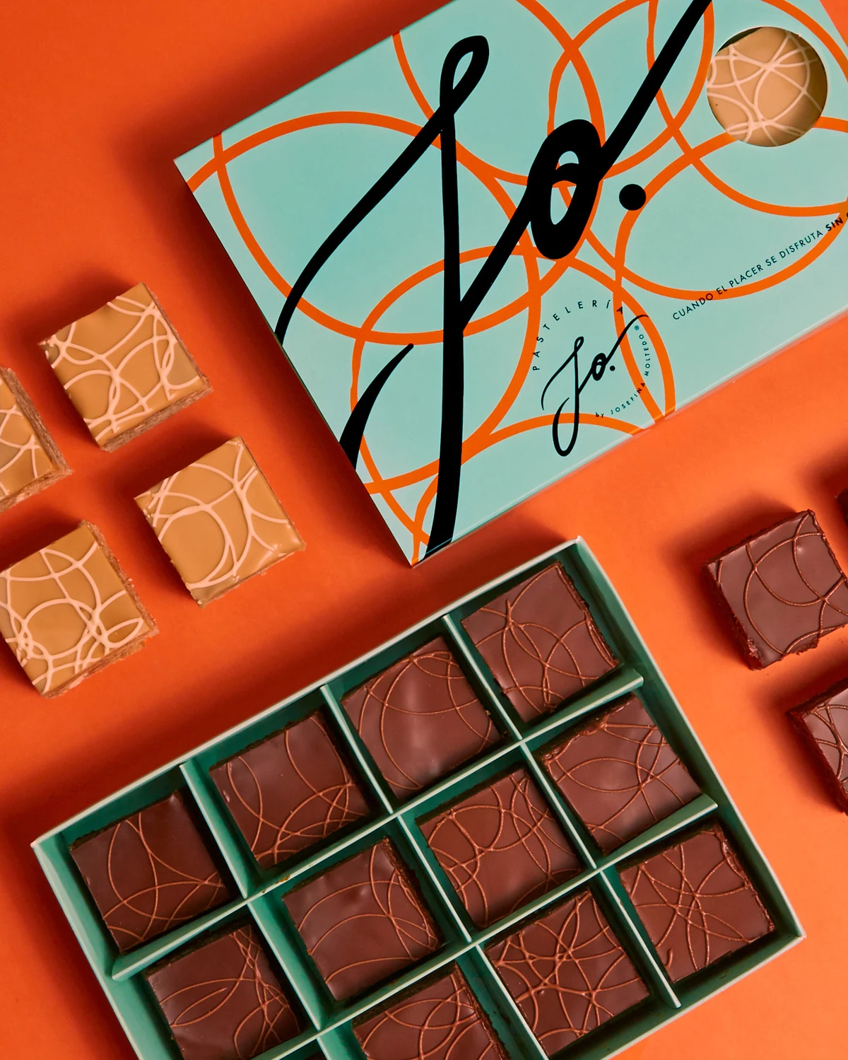 Caja JO en verde agua con la caligrafía de marca y patrón naranja, rodeada de cuadrados de chocolate y manjar sobre fondo naranja.