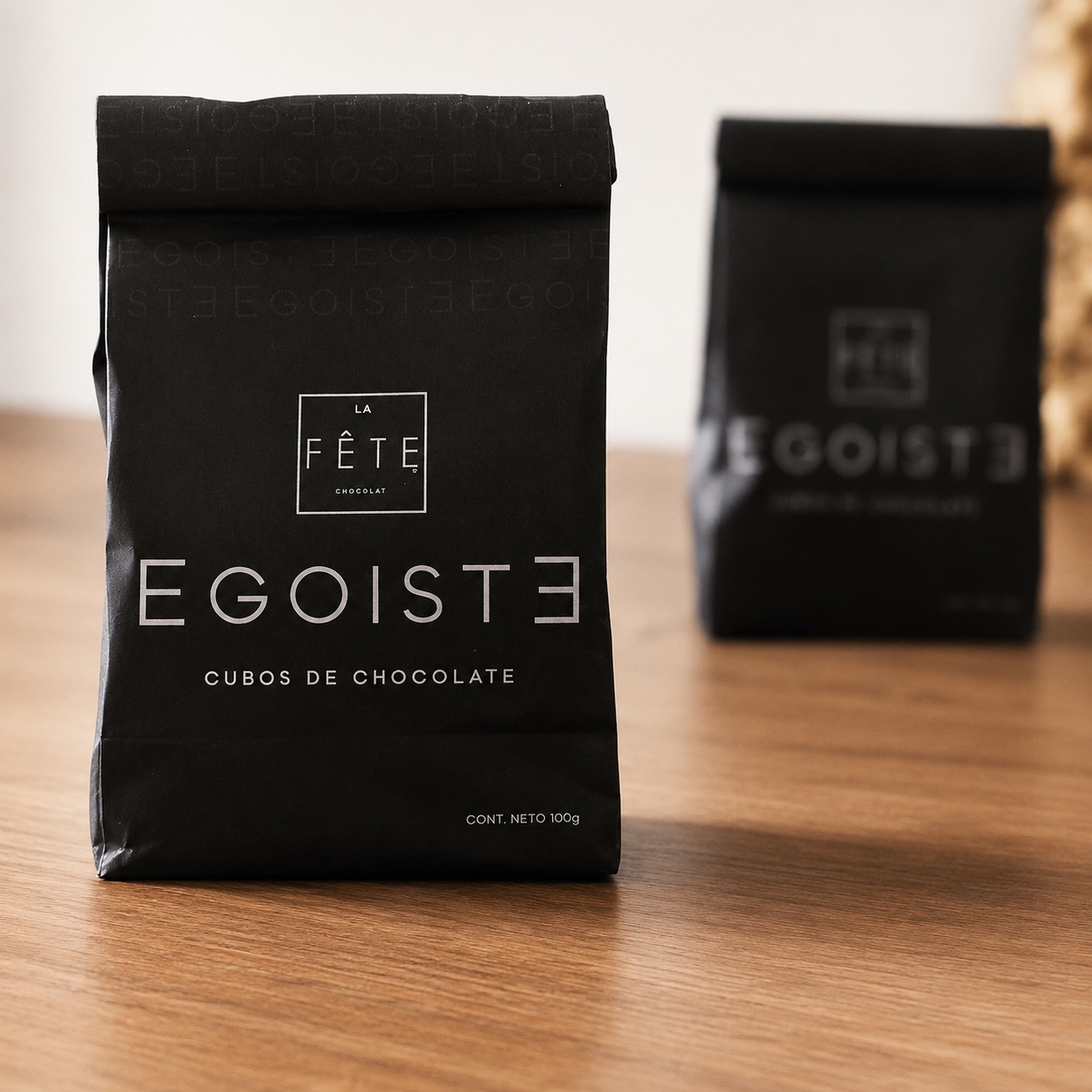 Bolsa negra de cubos de chocolate Egoíste de La Fête sobre una mesa de madera, con un segundo envase desenfocado al fondo