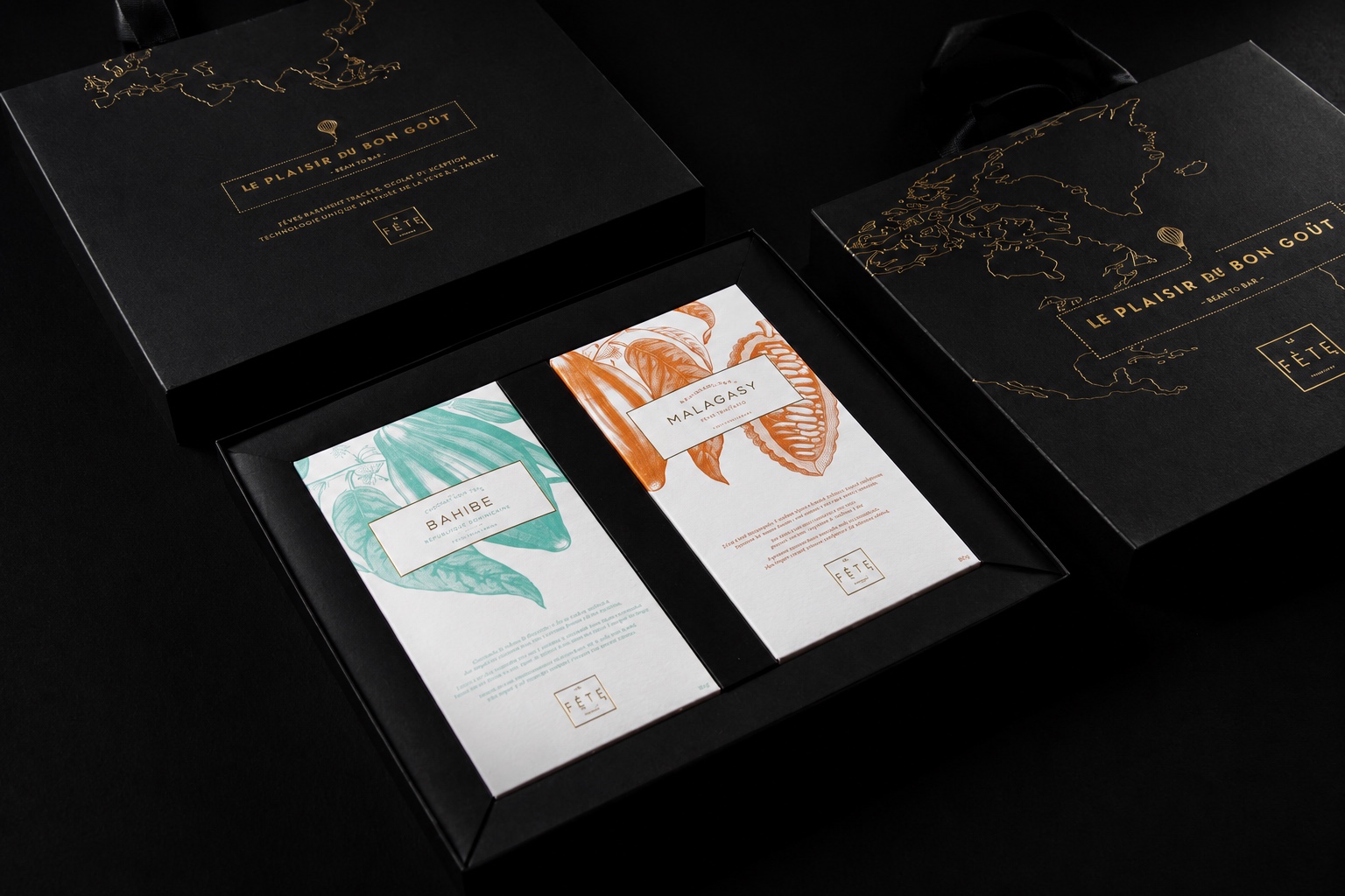 Estuche negro abierto de La Fête con dos barras de chocolate de origen Bahibe y Malagasy, rodeado de cajas negras con mapas en folia dorada