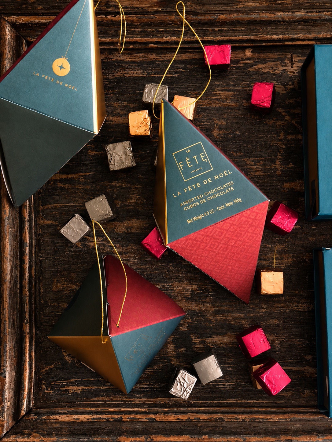 Estuches triangulares azul petróleo y rojo de La Fête de Noël, acompañados de chocolates envueltos sobre una bandeja de madera
