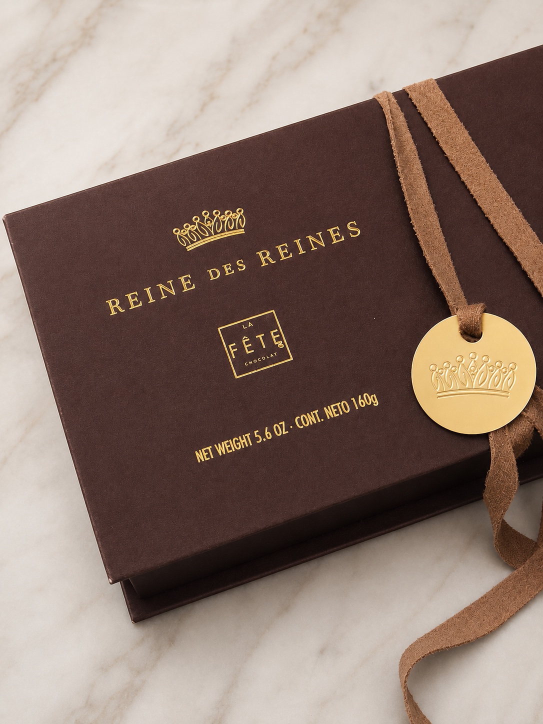 Estuche café Reine des Reines de La Fête con tipografía dorada, cinta tipo gamuza y medallón metálico sobre mármol claro
