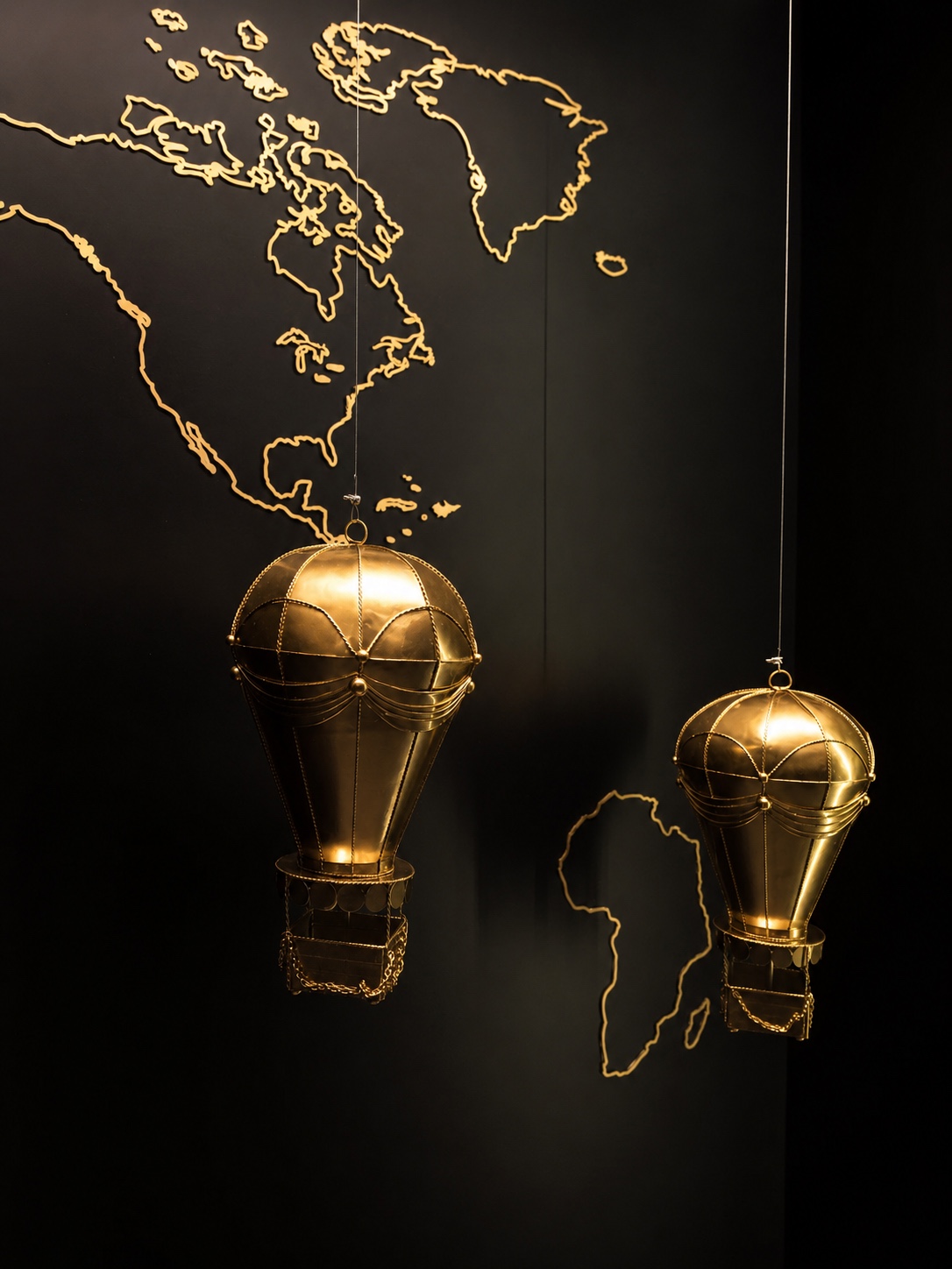 Dos globos aerostáticos metálicos dorados suspendidos frente a un mapa del mundo trazado en dorado sobre una pared negra