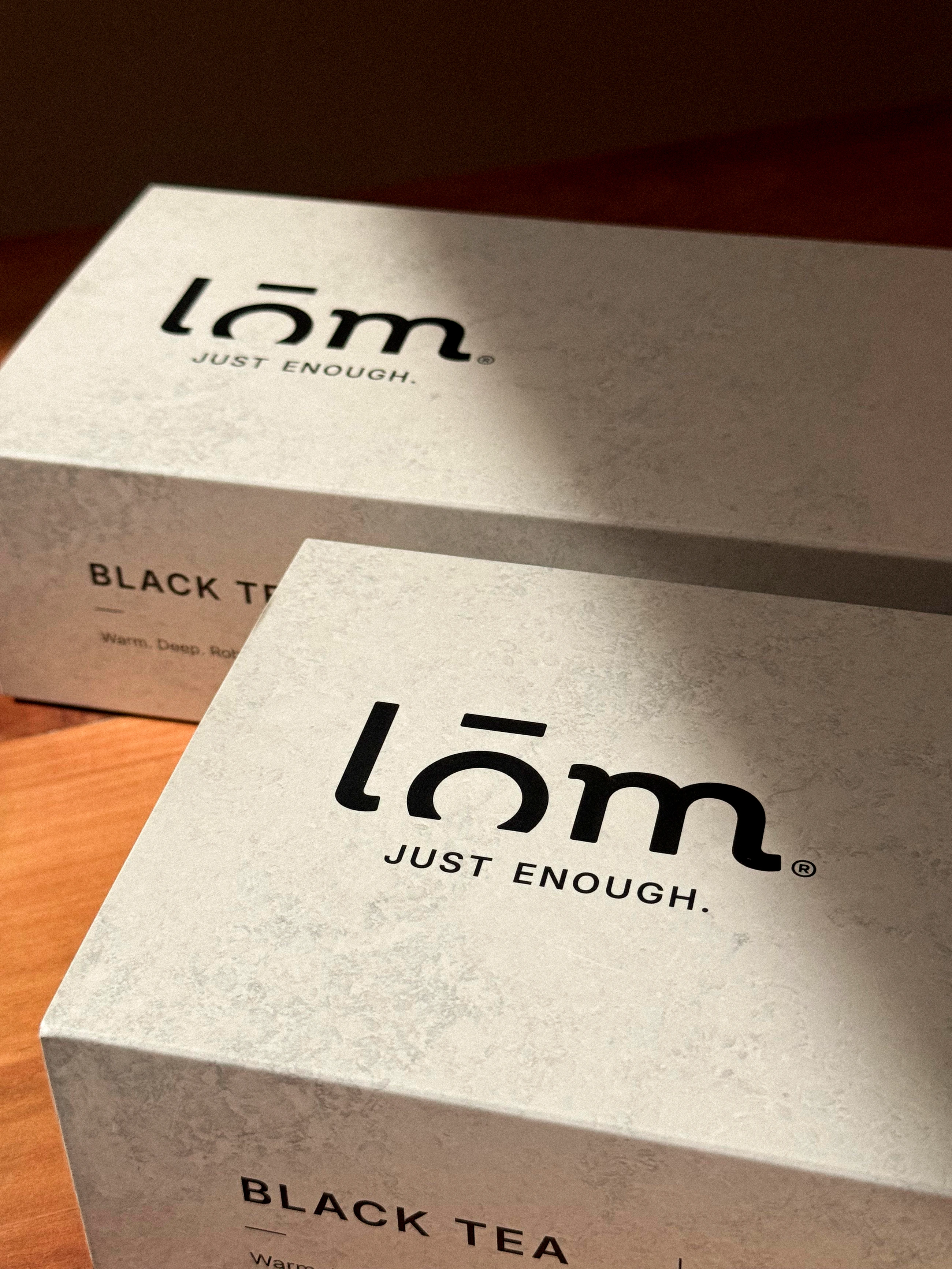 Dos cajas LŌM de té negro con logotipo negro sobre una superficie clara.