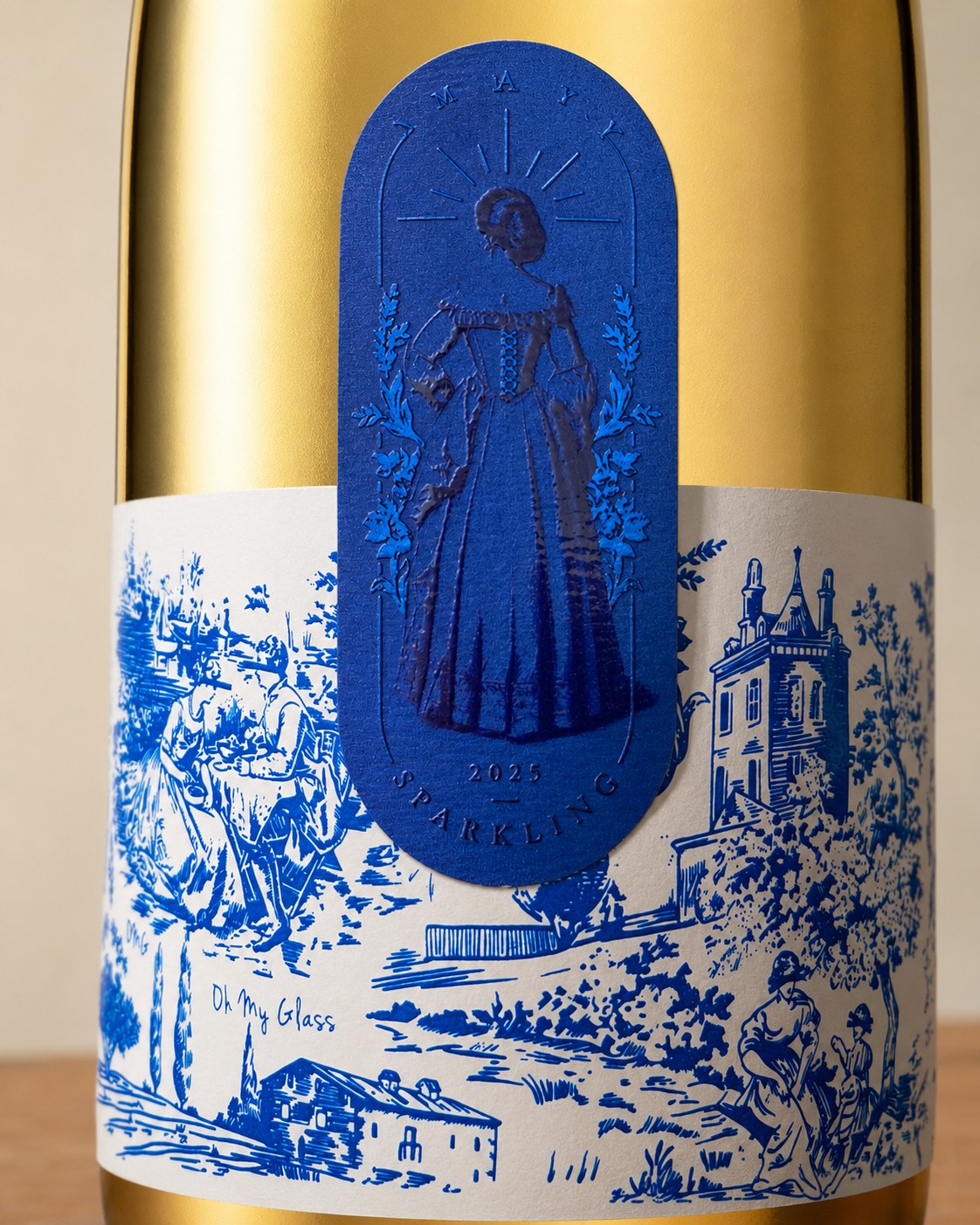 Detalle de etiqueta ilustrada estilo toile de jouy en azul sobre botella dorada Oh My Glass