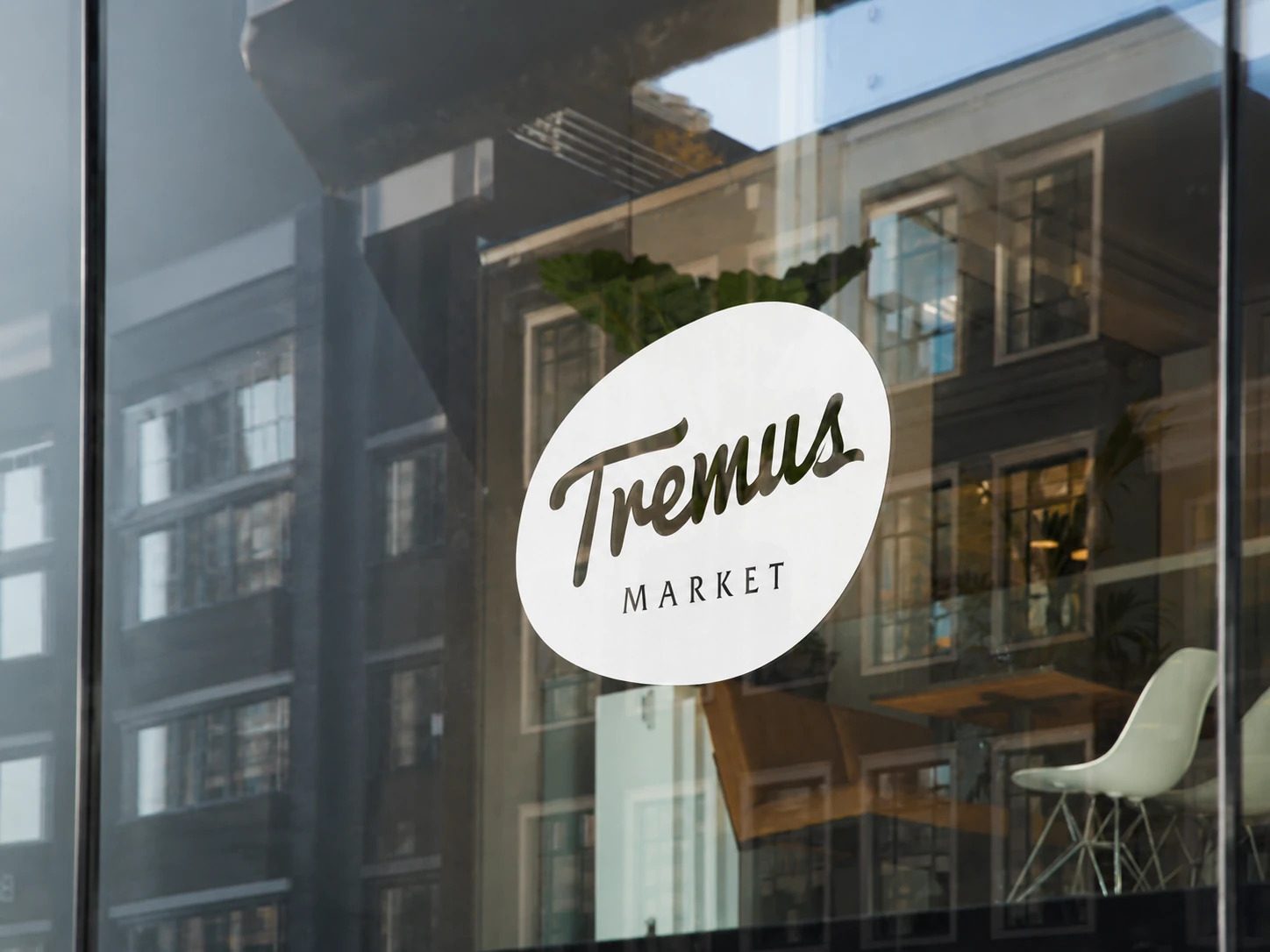 Isologo Tremus Market aplicado en vinilo blanco sobre el vidrio de una fachada urbana, con el edificio reflejado.