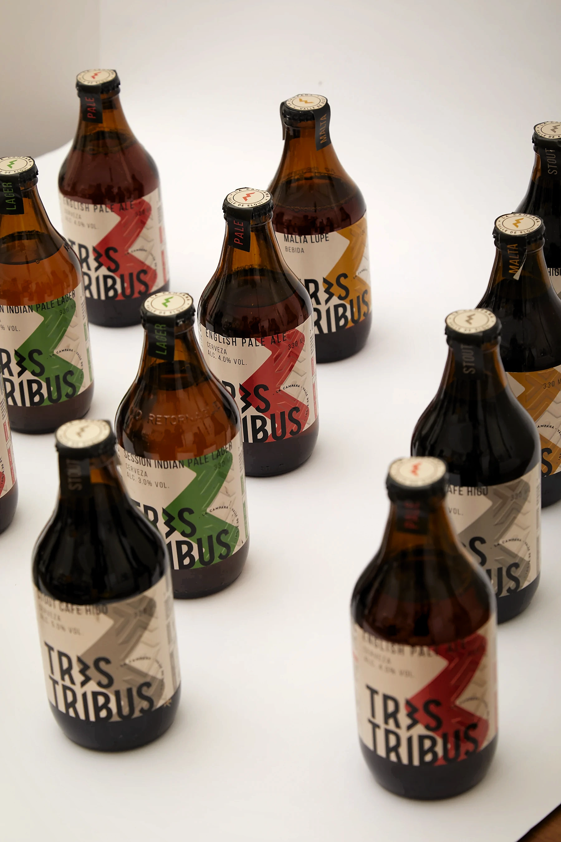 Botellas de cerveza Tres Tribus dispuestas en diagonal sobre fondo blanco, con etiquetas en papel crudo y zigzags rojo, ocre, gris y verde según la variedad.