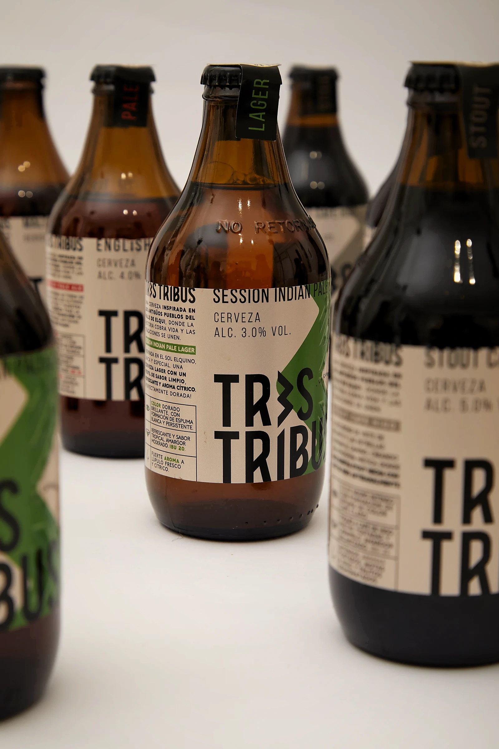Primer plano de la botella Session Indian Pale Lager de Tres Tribus, con zigzag verde sobre papel crudo y el collarín negro con la palabra LAGER.