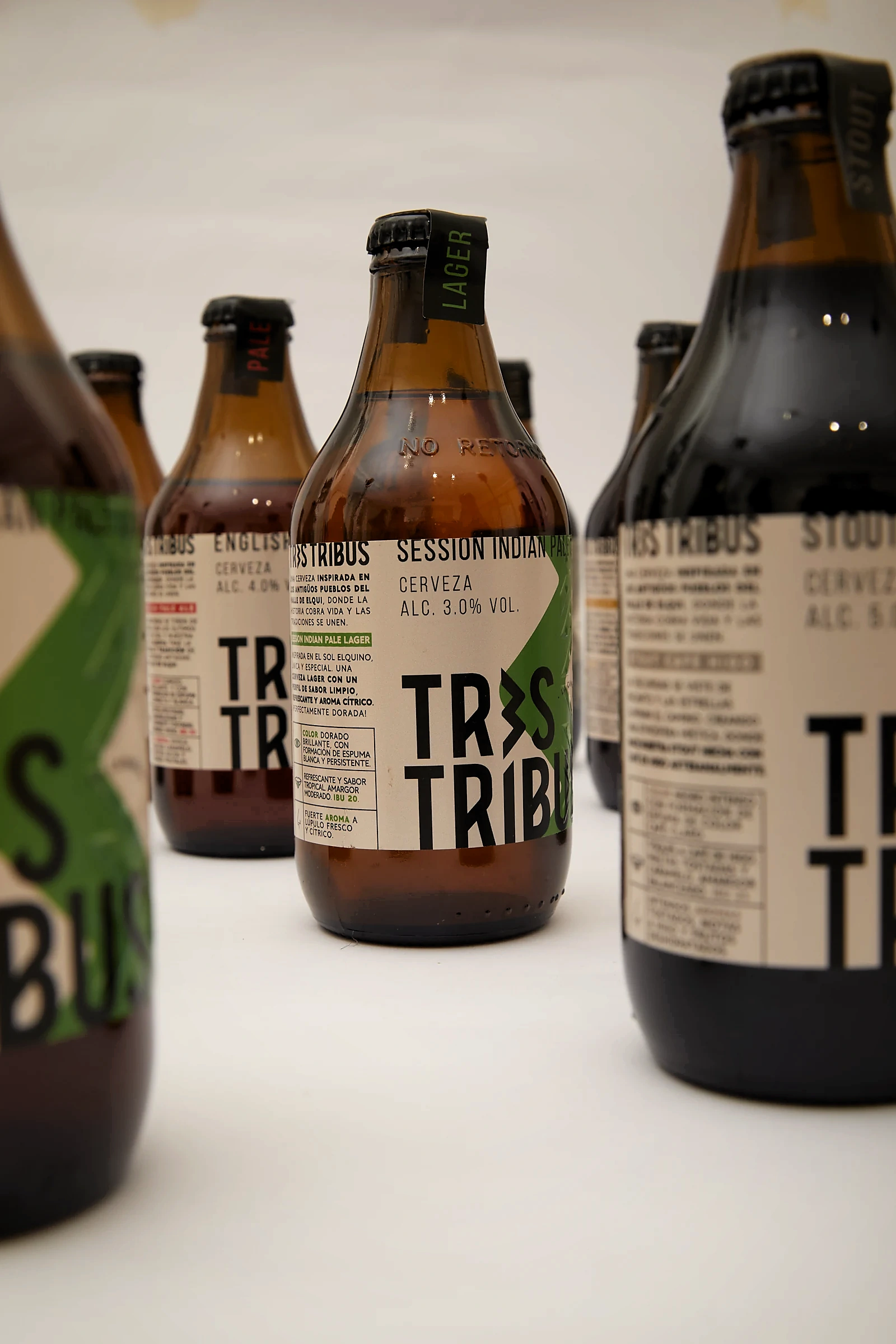 Varias botellas Tres Tribus en profundidad de campo: la Session Indian Pale Lager enfocada al centro y las variedades pale ale, malta y stout desenfocadas alrededor.
