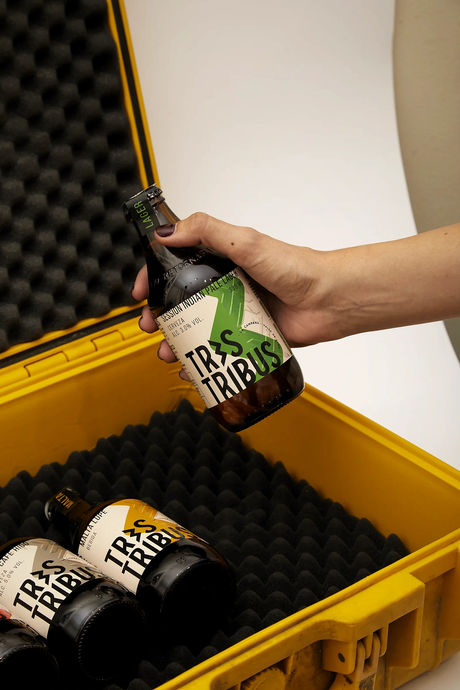 Una mano saca una botella Session Indian Pale Lager de un estuche rígido amarillo con espuma negra, donde quedan otras botellas Tres Tribus.
