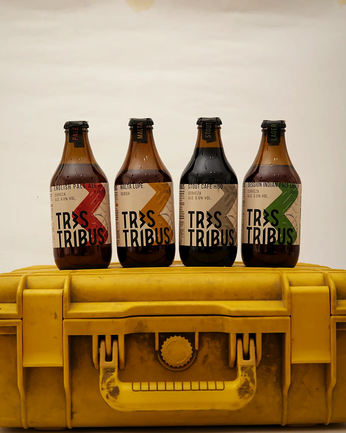 Cuatro botellas de cerveza Tres Tribus alineadas sobre un estuche rígido amarillo, con etiquetas en papel crudo y un zigzag rojo, ocre, gris y verde.