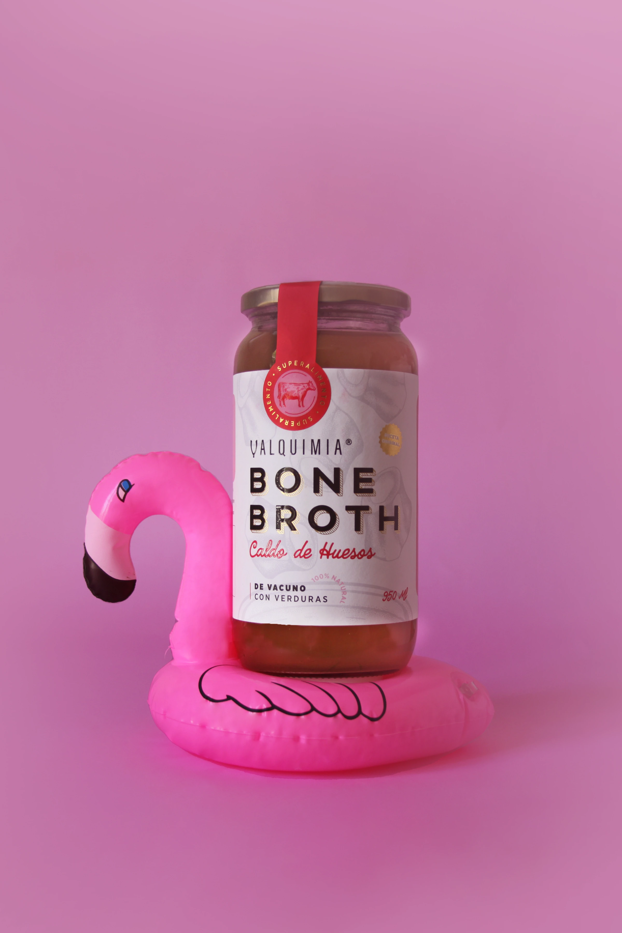 Frasco de Valquimia Bone Broth apoyado sobre un flamenco rosado, en un fondo rosa.
