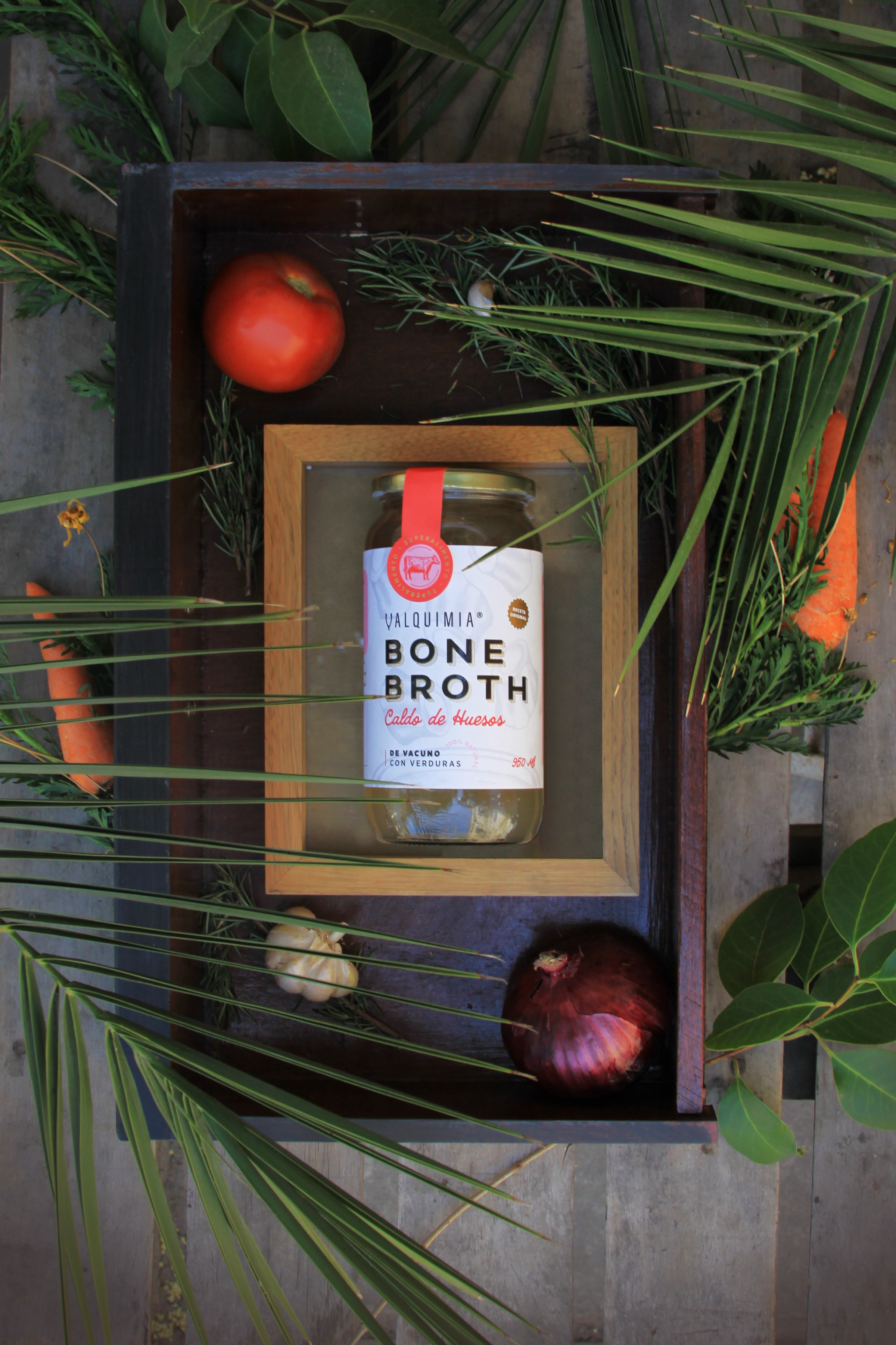 Frasco de Valquimia Bone Broth contenido en una caja de madera, rodeado por verduras y follaje.