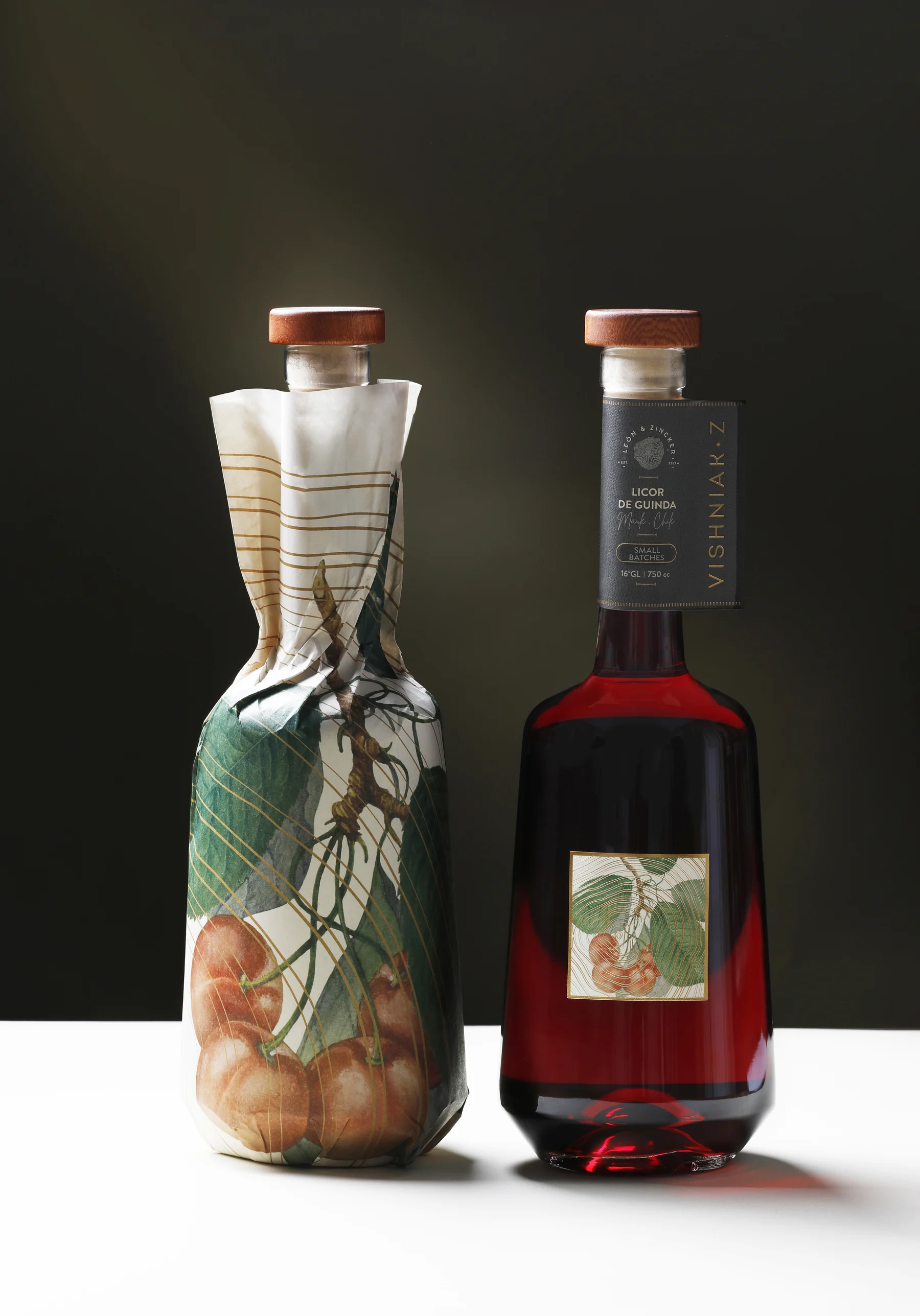 Dos botellas de Vishniak Z lado a lado: una envuelta en el papel protector ilustrado con guindas y raíces, la otra desnuda mostrando el licor rojo rubí y el colgante de cuello.
