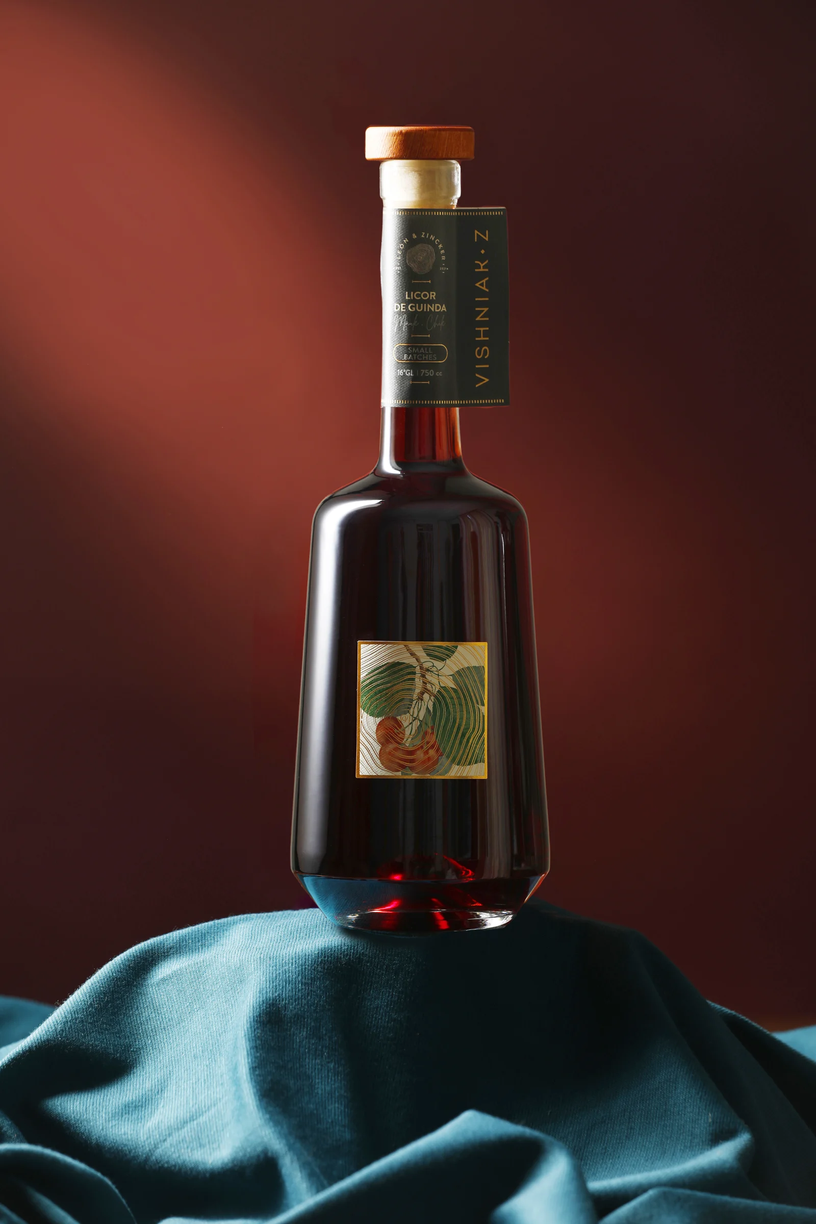 Retrato vertical completo de la botella Vishniak Z sobre tela azul petróleo, con fondo granate y la etiqueta ilustrada de guindas al centro.