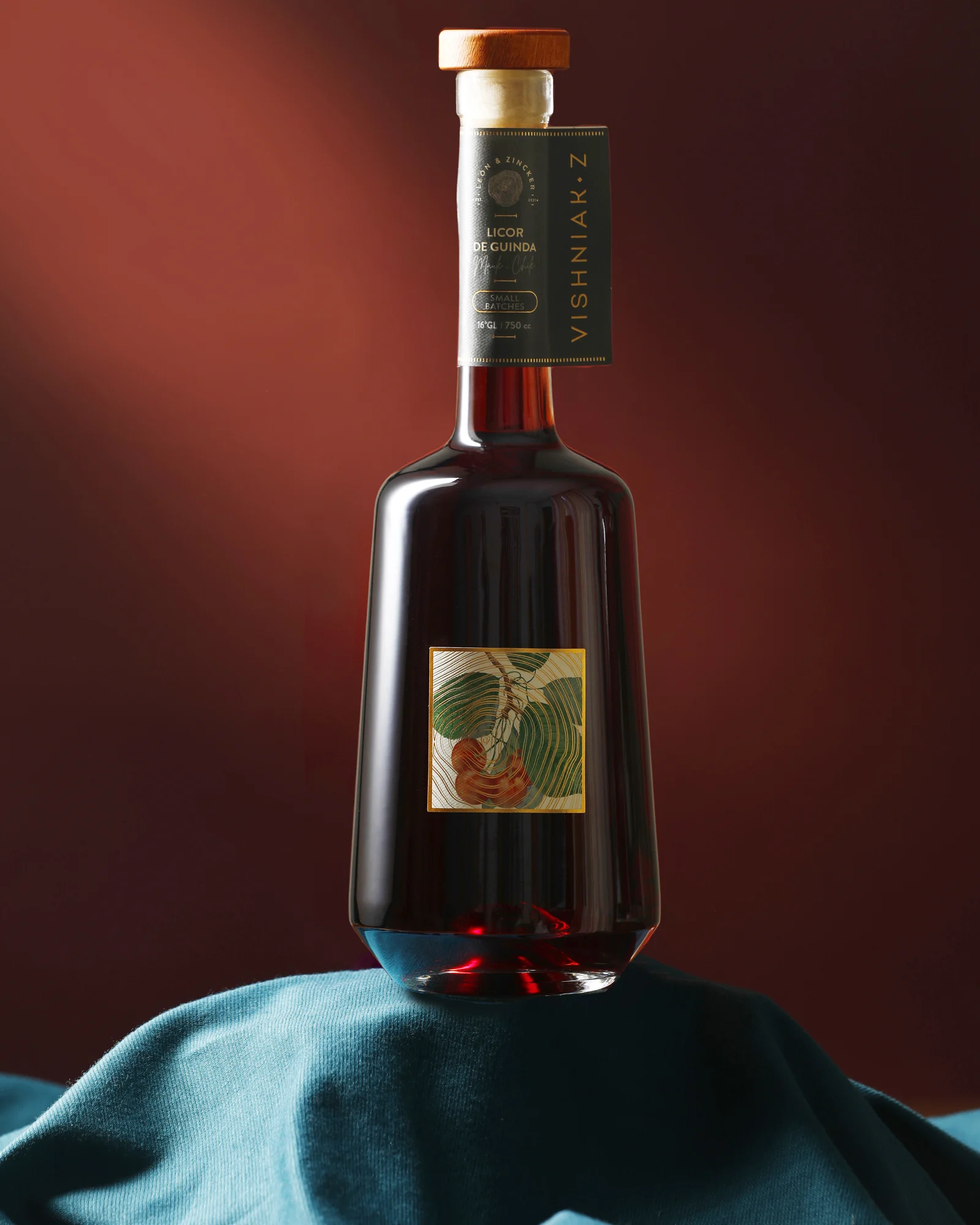 Botella de Vishniak Z sobre una tela azul petróleo, con etiqueta ilustrada de guindas y colgante de cuello sobre fondo granate.