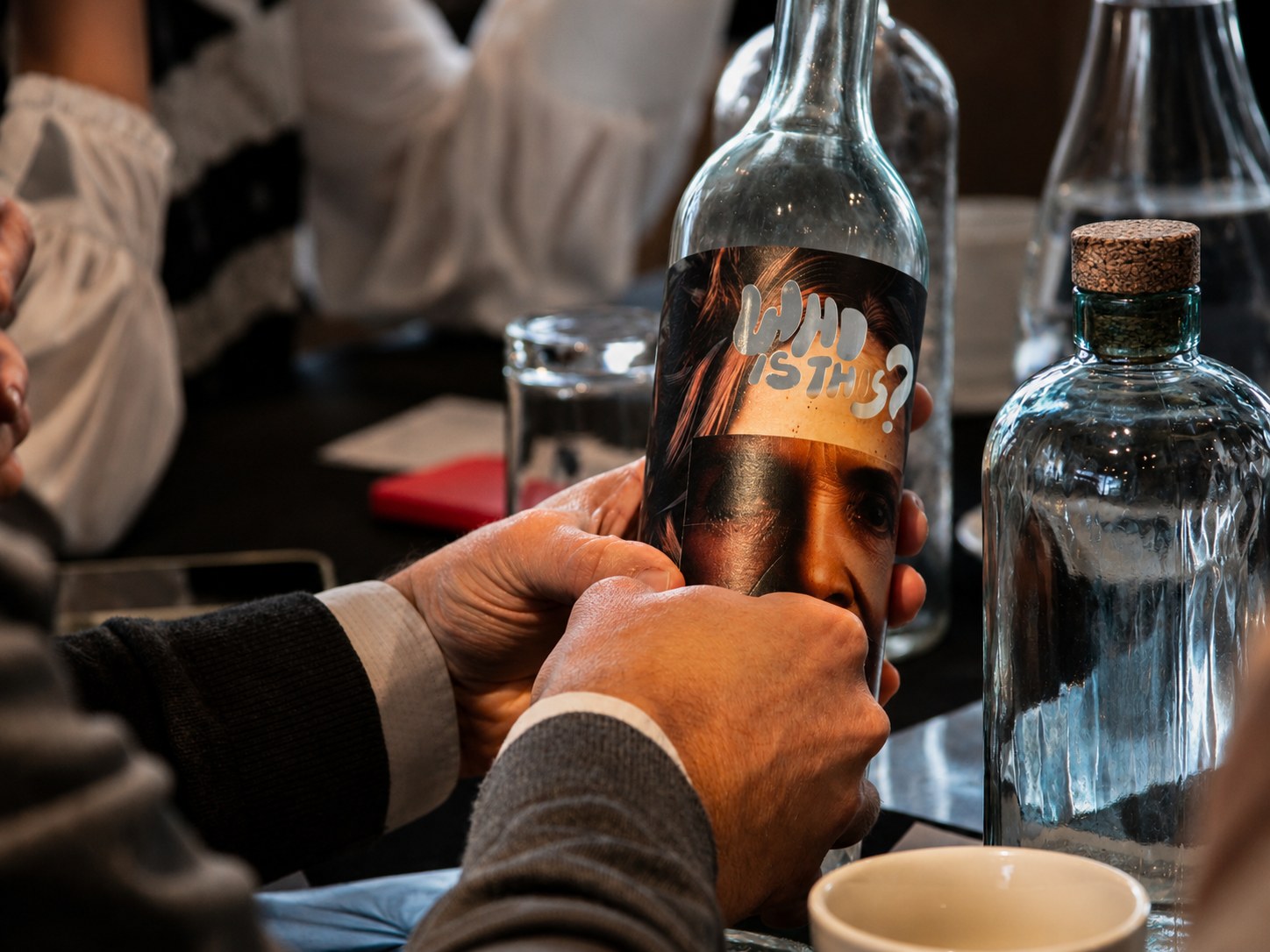 Manos aplicando una etiqueta con un rostro y el texto WHO IS THIS sobre una botella de vidrio durante el taller de intervención