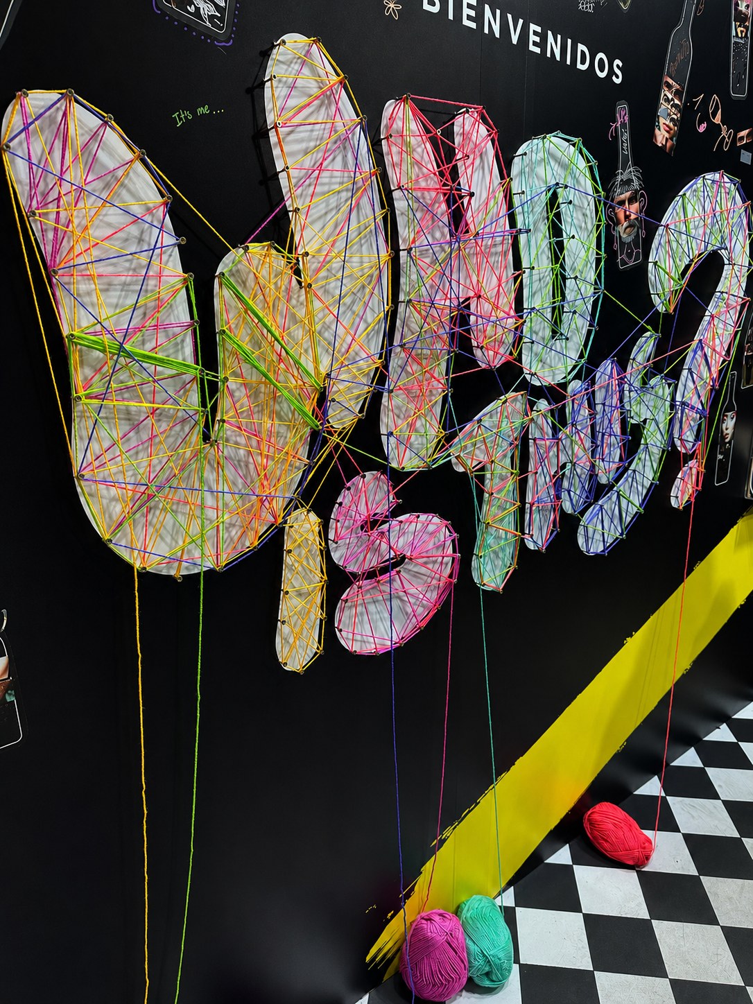 Instalación de hilos de colores formando el texto WHO IS THIS sobre un fondo a cuadros