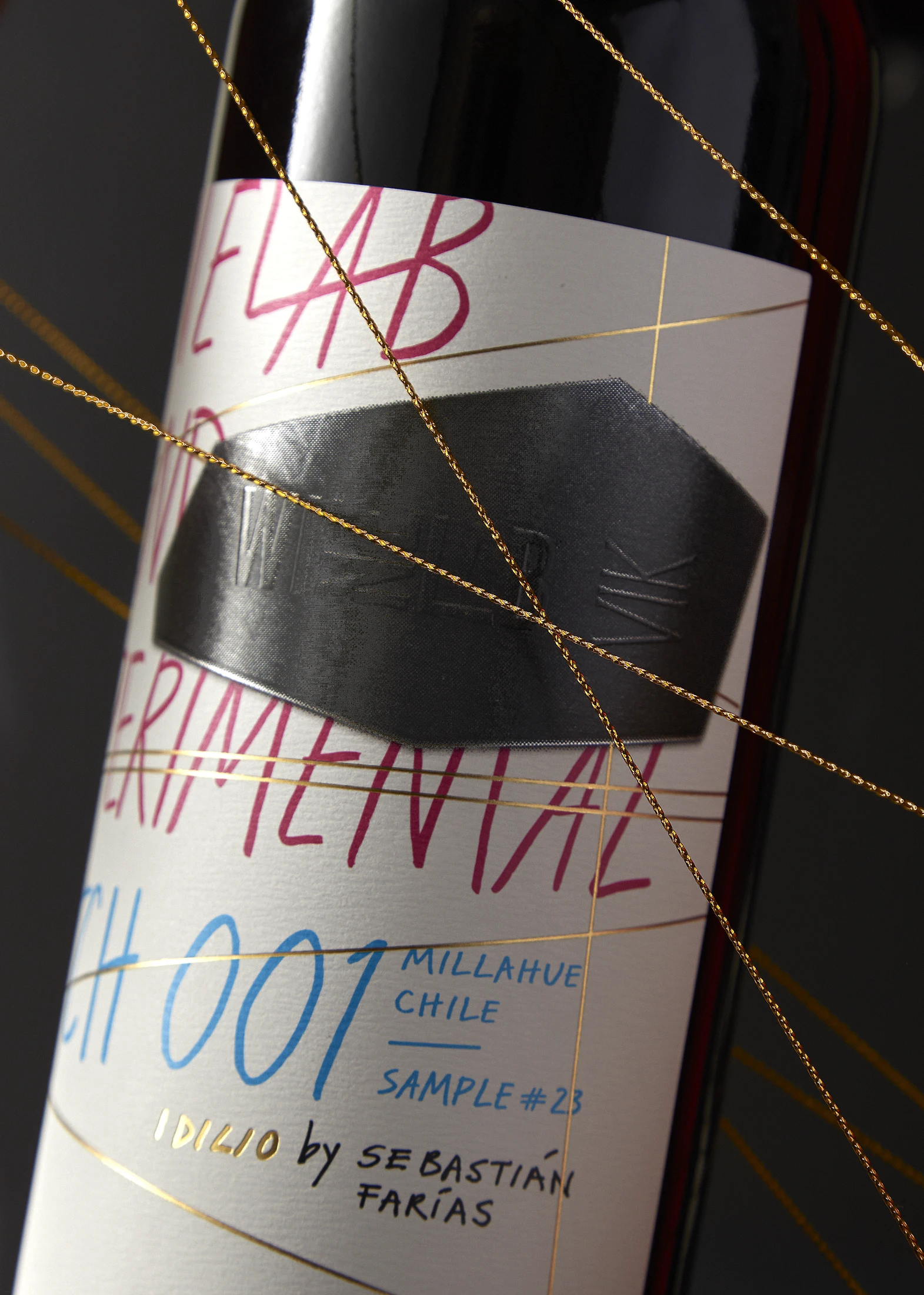Detalle de la etiqueta WineLab Experimental Batch 001: escritura a mano en magenta y azul, placa metálica en relieve e hilo dorado cruzando la botella.