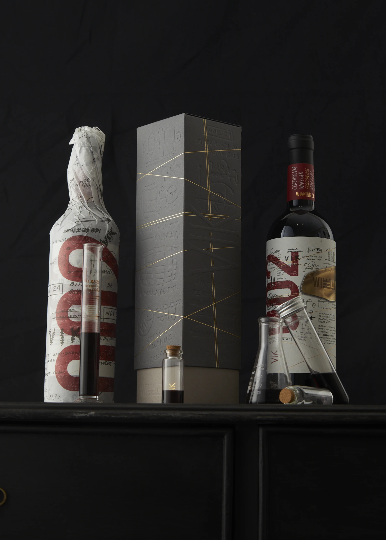 Edición 2024 de VIK WineLab: la botella envuelta en papel anotado con el numeral 002, el estuche gris con relieve y líneas de foil, y la botella final con su etiqueta escrita a mano.