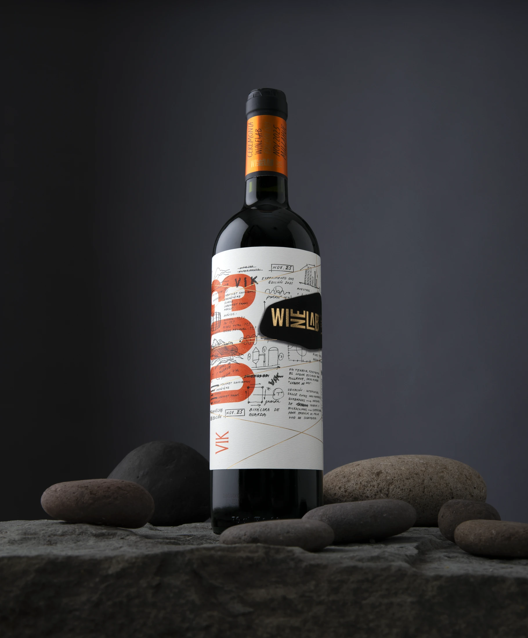Botella WineLab 2025 de pie sobre piedras, con cápsula naranja y la etiqueta de bitácora de guarda.