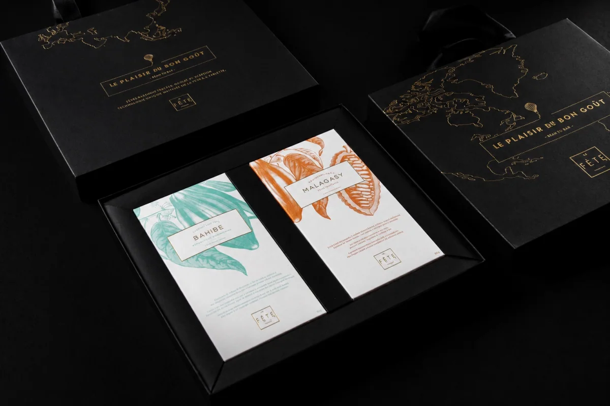 Estuche negro abierto de La Fête con dos barras de chocolate de origen Bahibe y Malagasy, rodeado de cajas negras con mapas en folia dorada