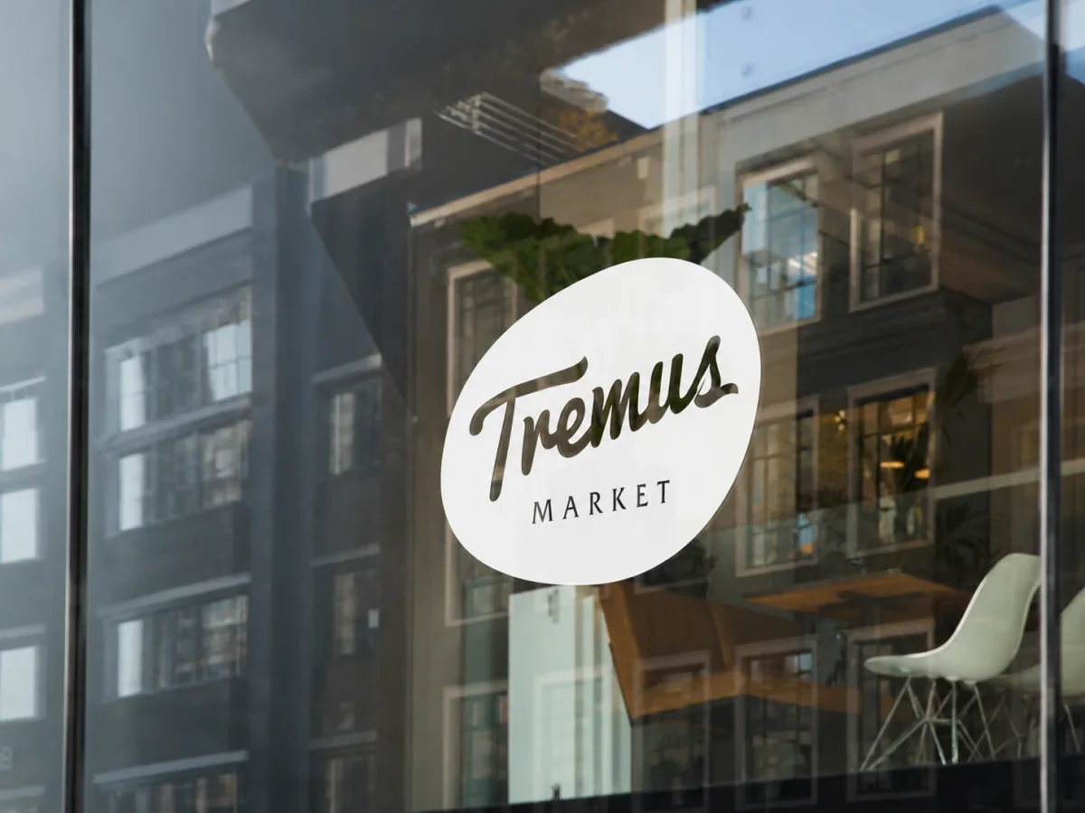 Isologo Tremus Market aplicado en vinilo blanco sobre el vidrio de una fachada urbana, con el edificio reflejado.