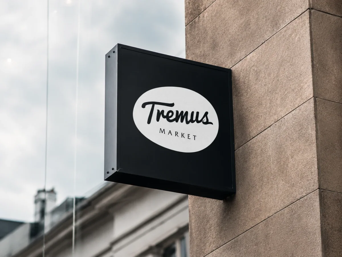 Letrero de fachada Tremus Market en caja negra con isologo blanco, montado sobre un muro de piedra.