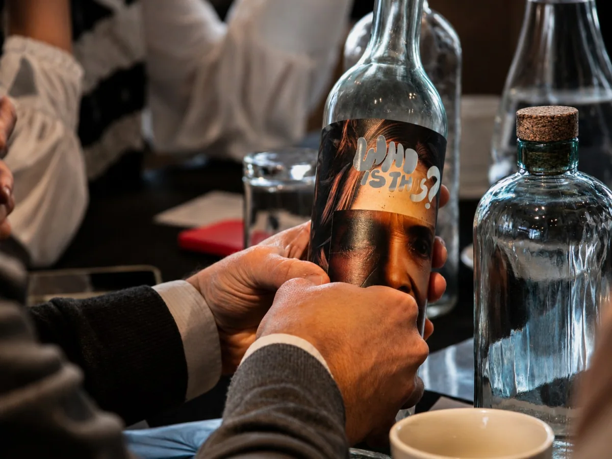 Manos aplicando una etiqueta con un rostro y el texto WHO IS THIS sobre una botella de vidrio durante el taller de intervención