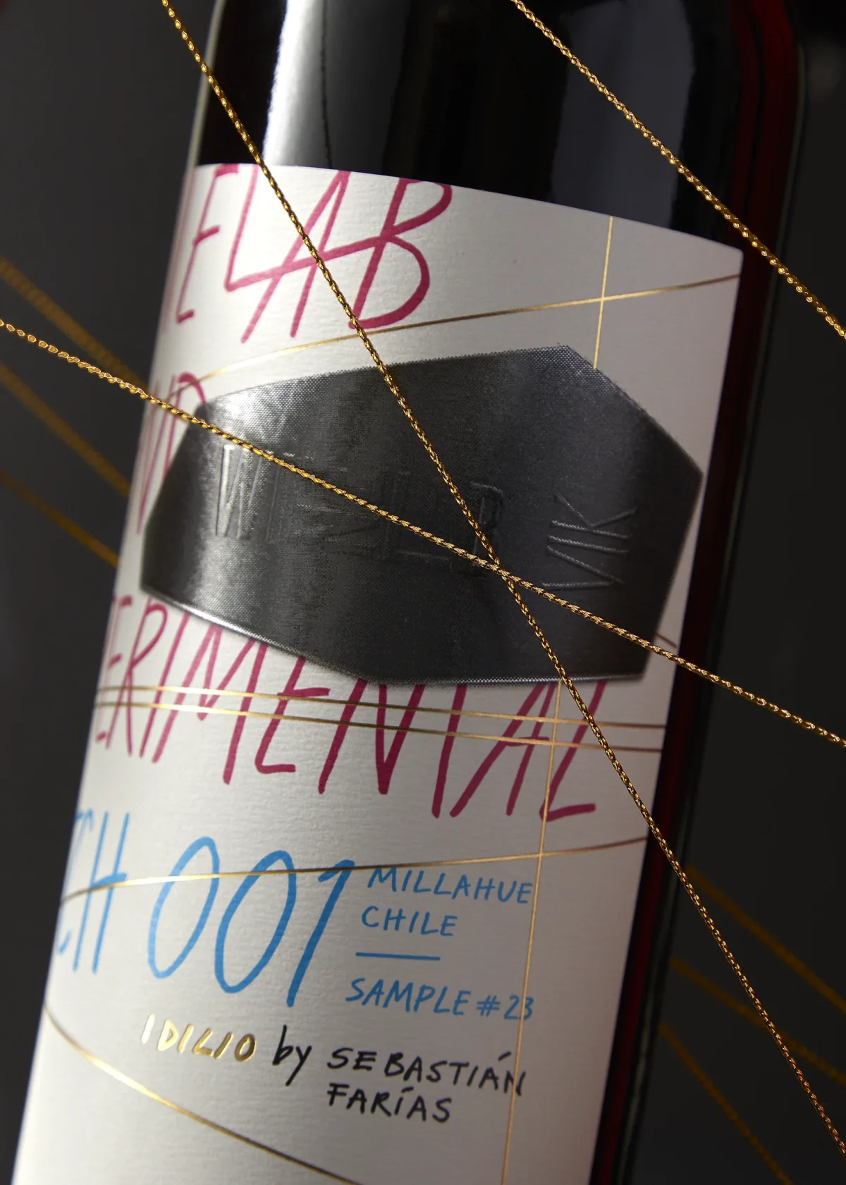Detalle de la etiqueta WineLab Experimental Batch 001: escritura a mano en magenta y azul, placa metálica en relieve e hilo dorado cruzando la botella.