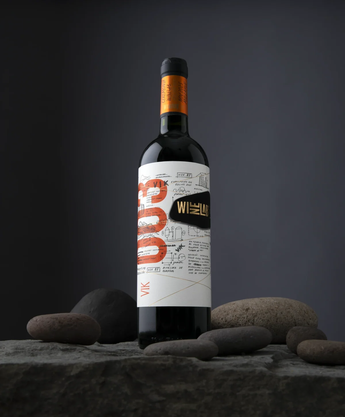 Botella WineLab 2025 de pie sobre piedras, con cápsula naranja y la etiqueta de bitácora de guarda.